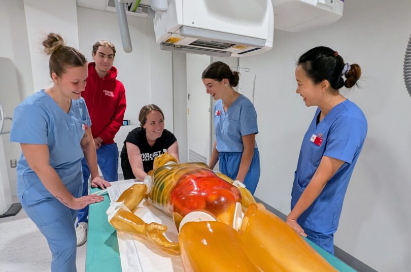 Students in scrubs gather around a medical mannequin in a radiology lab during a hands-on learning activity.