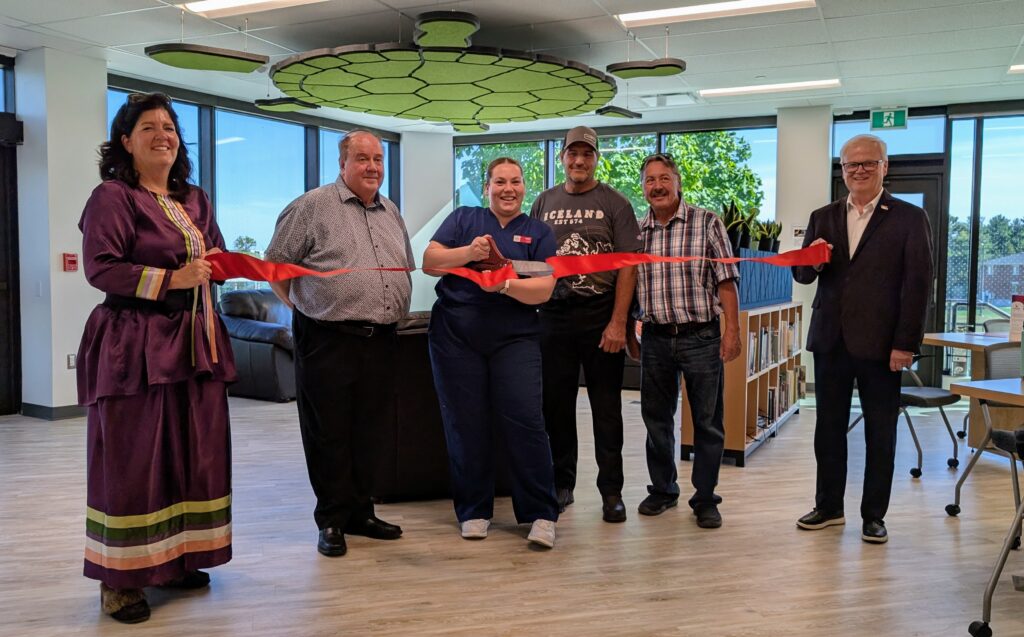 Six people smile while cutting a red ribbon during an opening ceremony inside a bright campus space.