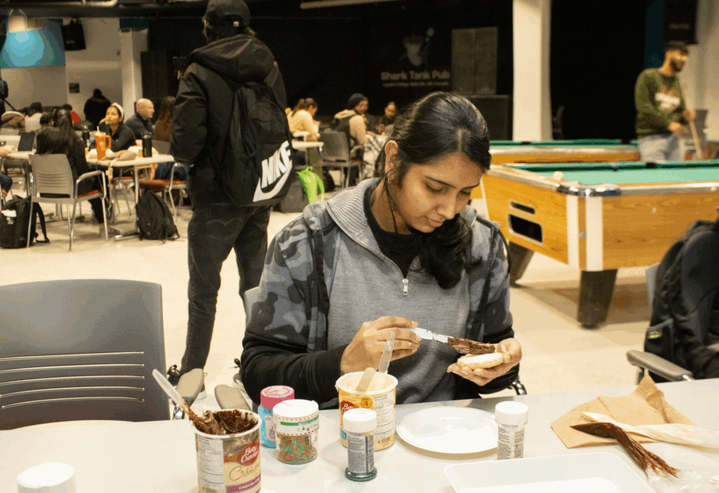A student decorates a cookie with chocolate icing at a table filled with baking supplies.