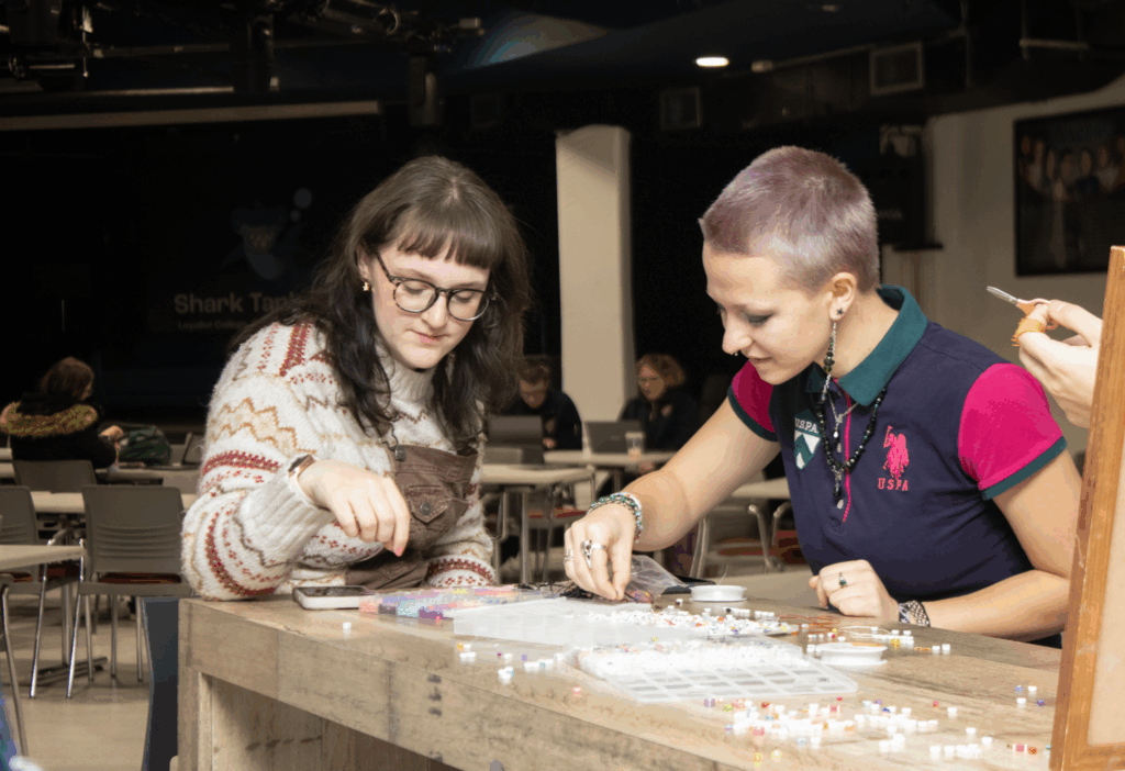 Two students sit at a table creating beaded crafts together during a campus activity.