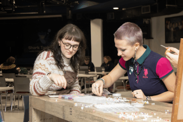 Two students sit at a table creating beaded crafts together during a campus activity.