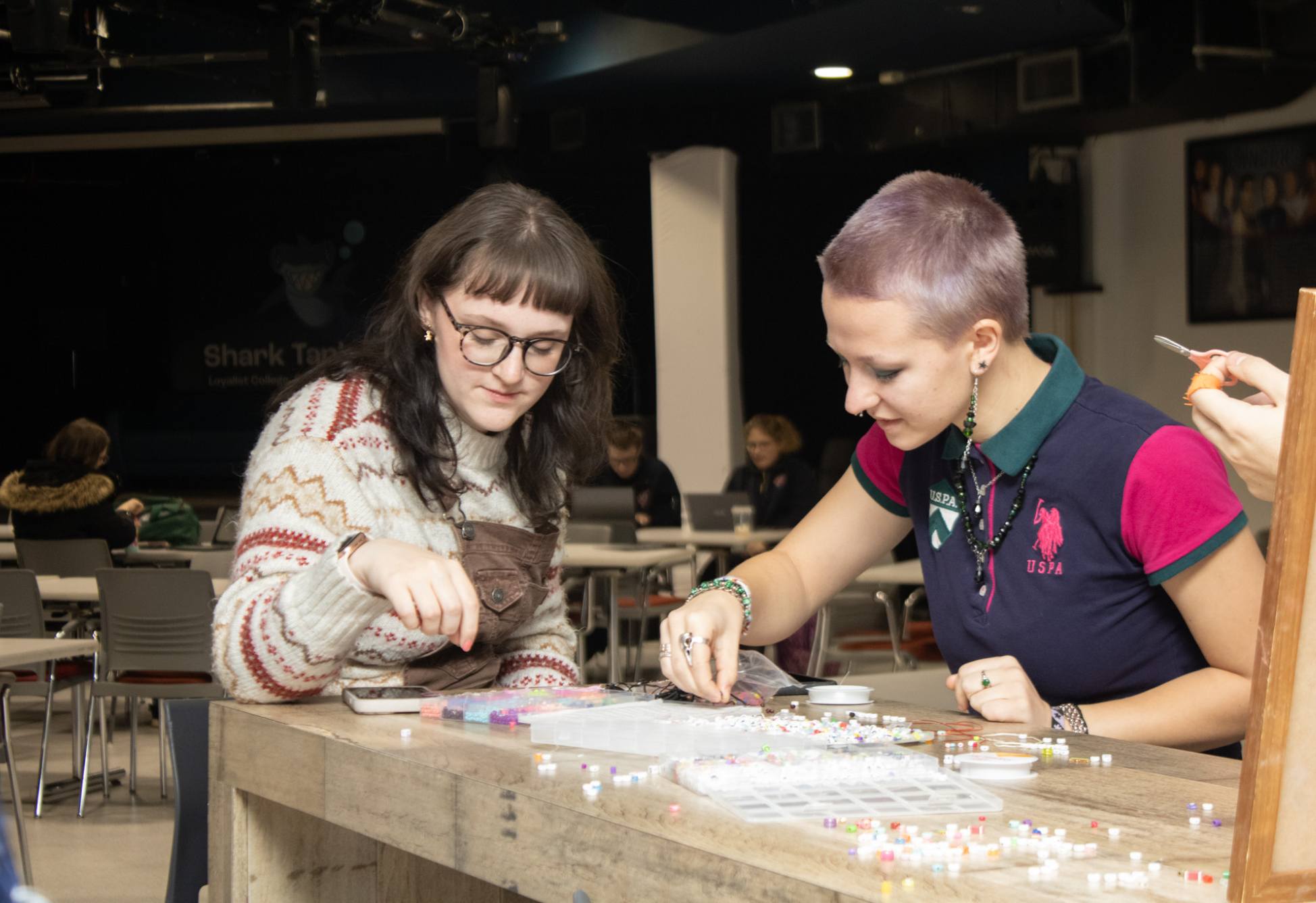 Two students sit at a table creating beaded crafts together during a campus activity.