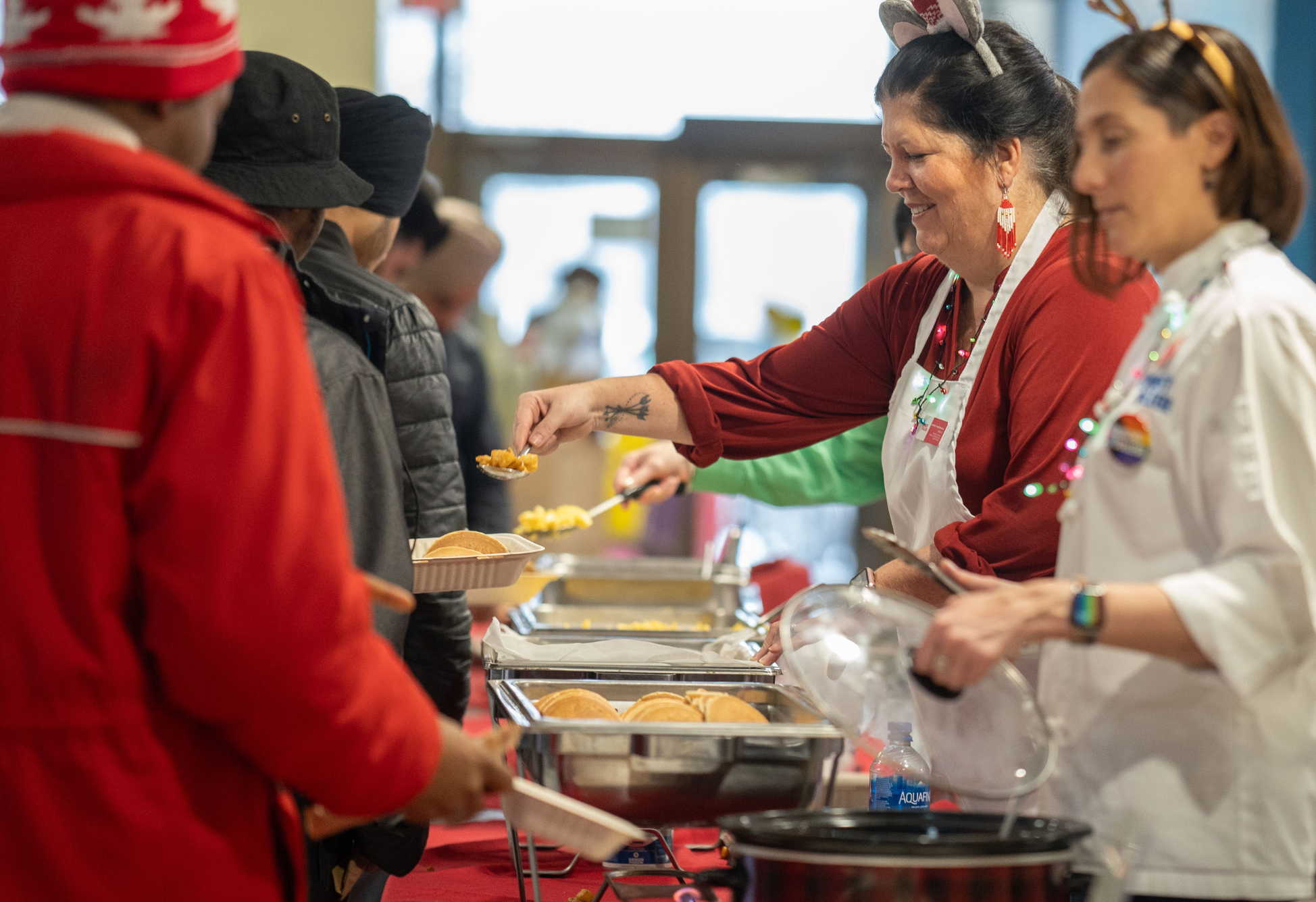 Staff wearing holiday attire serve food to students from buffet trays during a festive meal event.