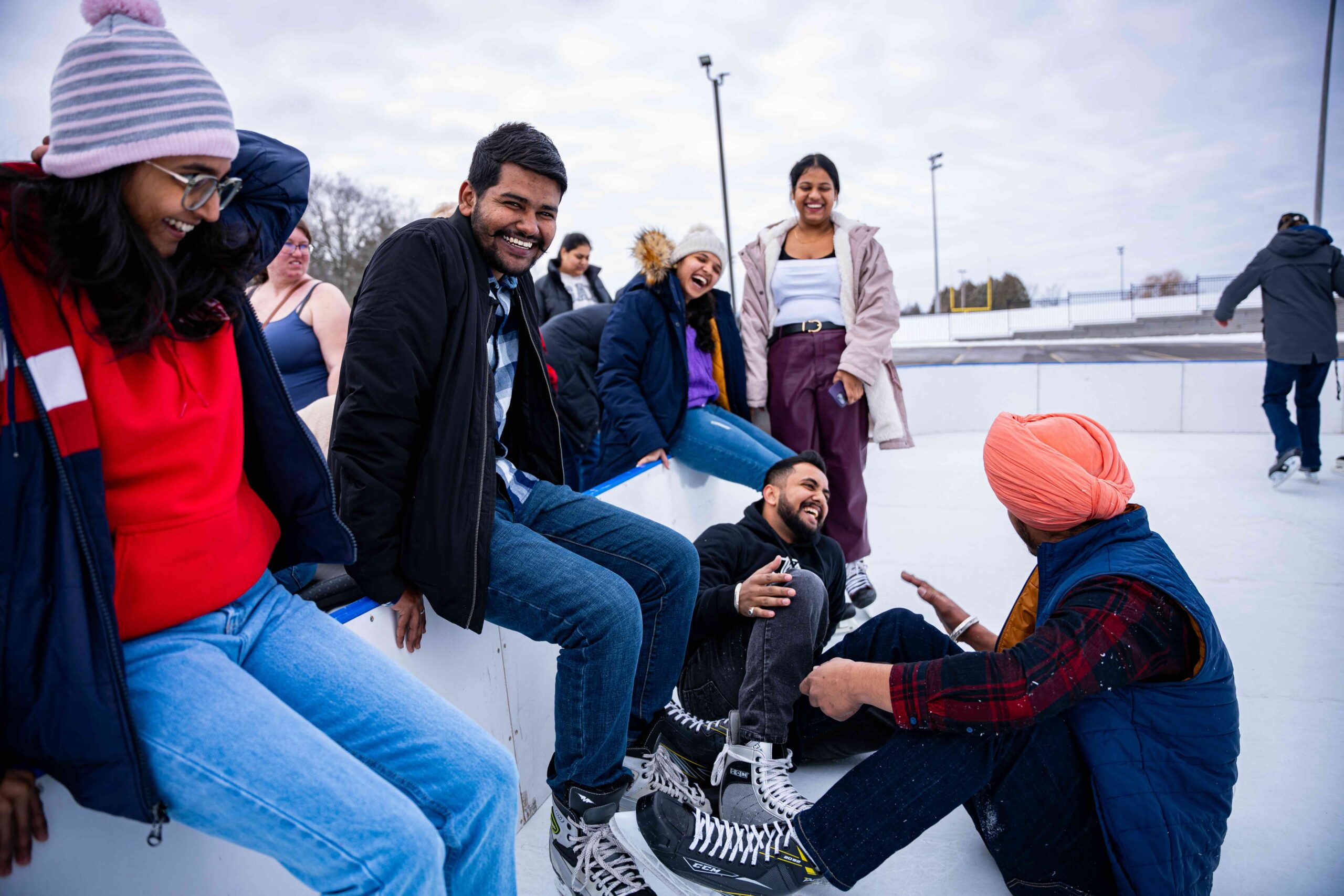 A group of students laugh and enjoy themselves at an outdoor skating rink. Some are sitting along the rink boards while others stand nearby, bundled in winter jackets, hats, and scarves. Two students wearing skates sit on the ice, smiling and talking.