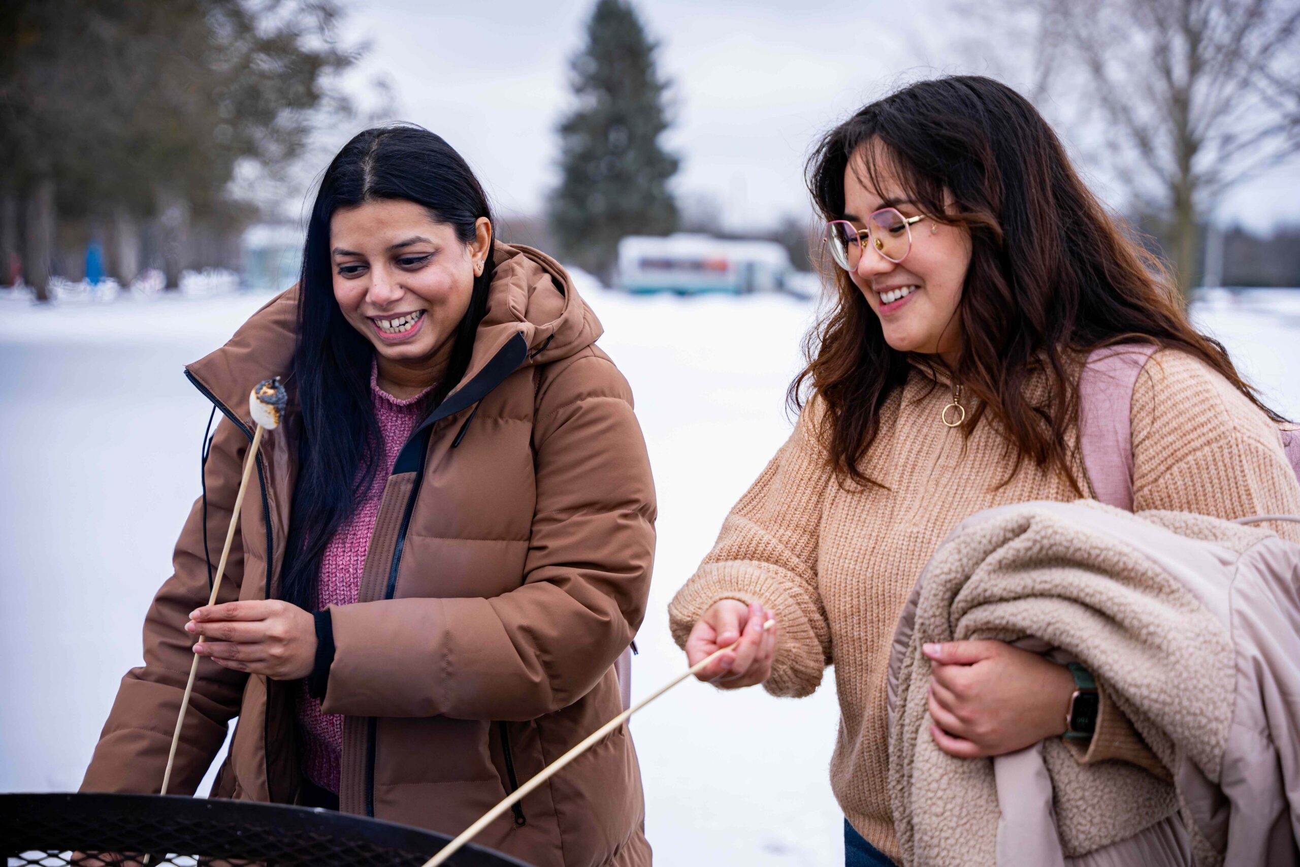 Two students smile as they roast marshmallows over an outdoor fire pit on a snowy winter day. One holds a toasted marshmallow on a skewer, while the other holds a blanket and her skewer, enjoying the moment together. Snow-covered trees and a blurred campus background sit behind them.