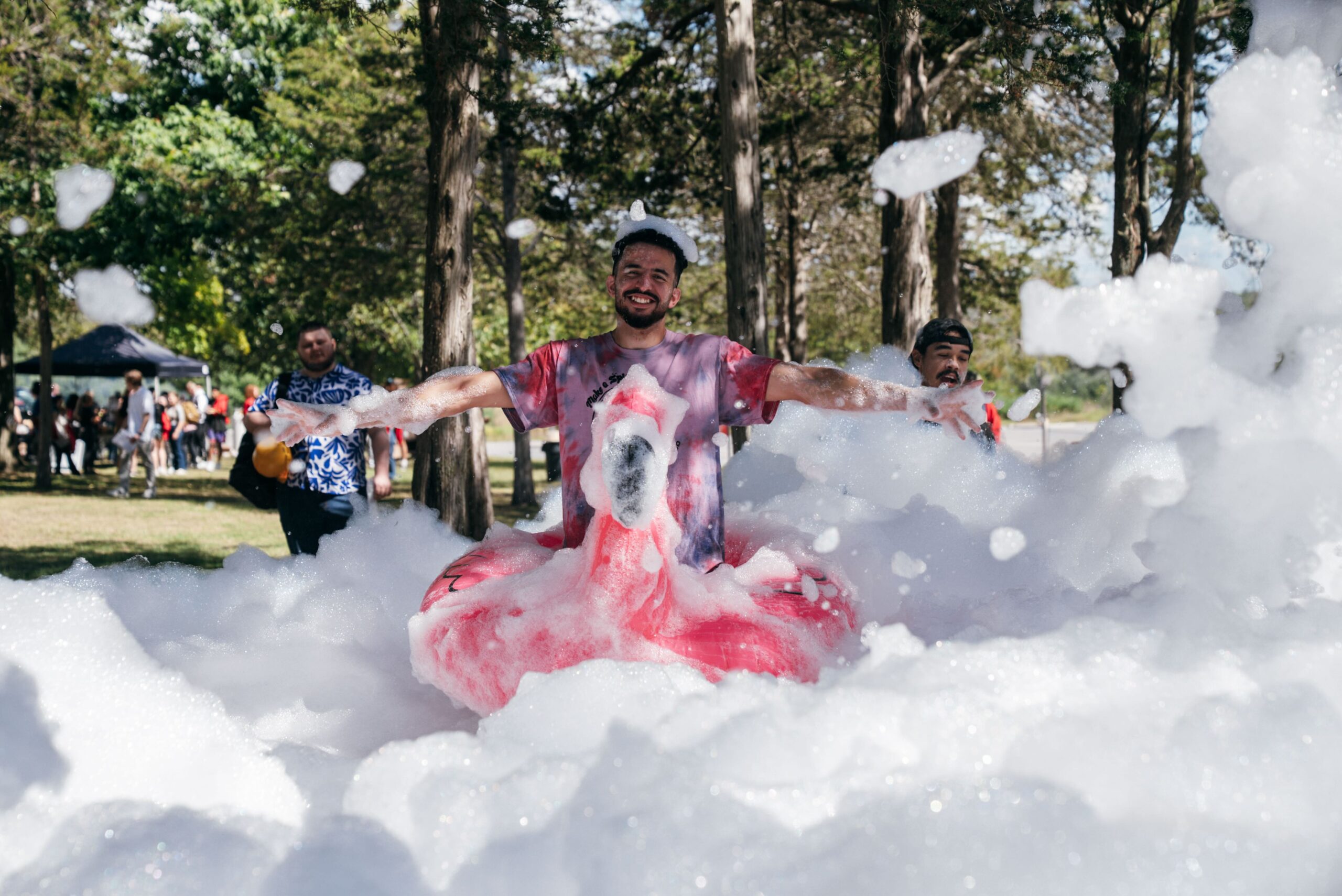 A student smiles and spreads their arms while sitting on a large pink flamingo float surrounded by thick white foam at an outdoor foam party during Orientation. Trees and other students can be seen in the background as bubbles fill the scene.