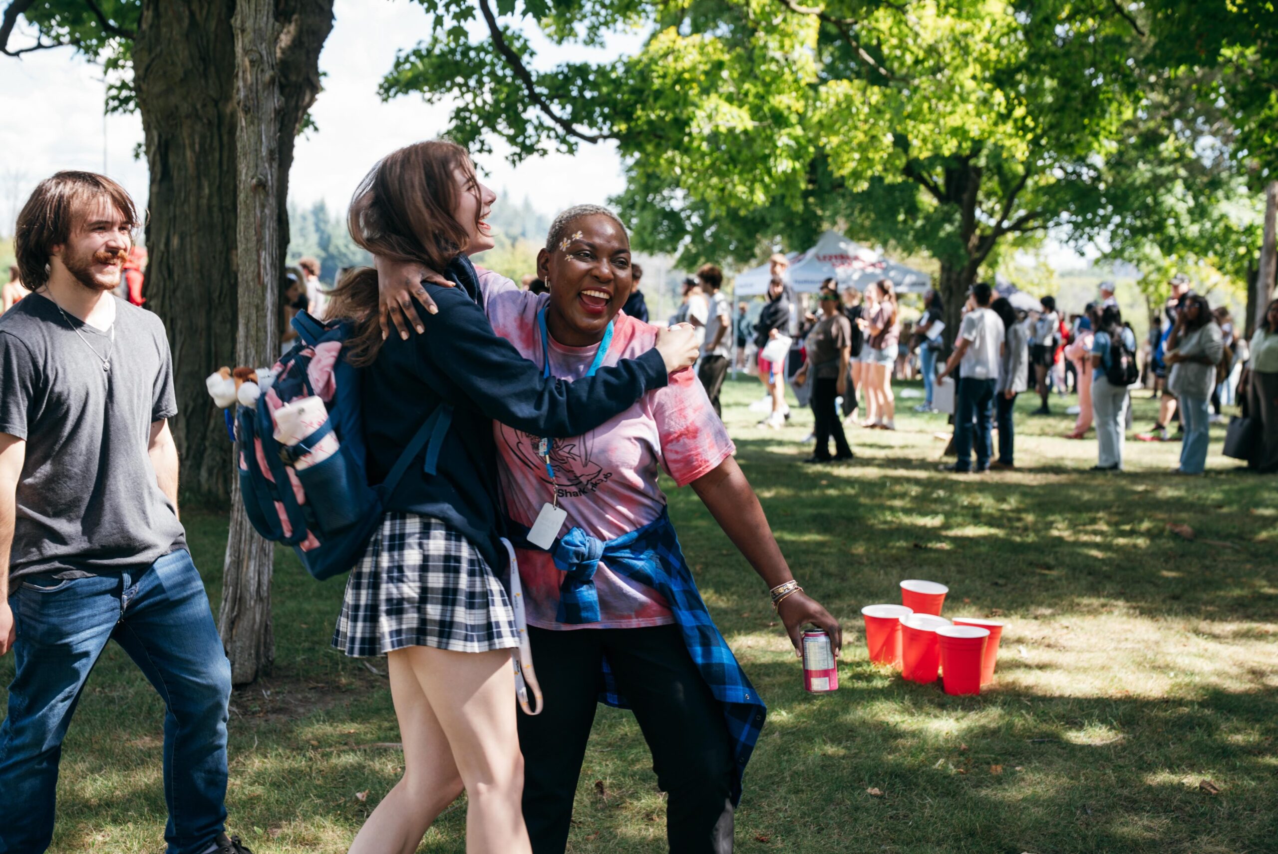 Two students laugh and hug during an outdoor Orientation event on a sunny day. One holds a drink can while the other carries a backpack. Other students gather and socialize in the background under the trees, with red plastic cups set up on the grass nearby for a game of water pong.