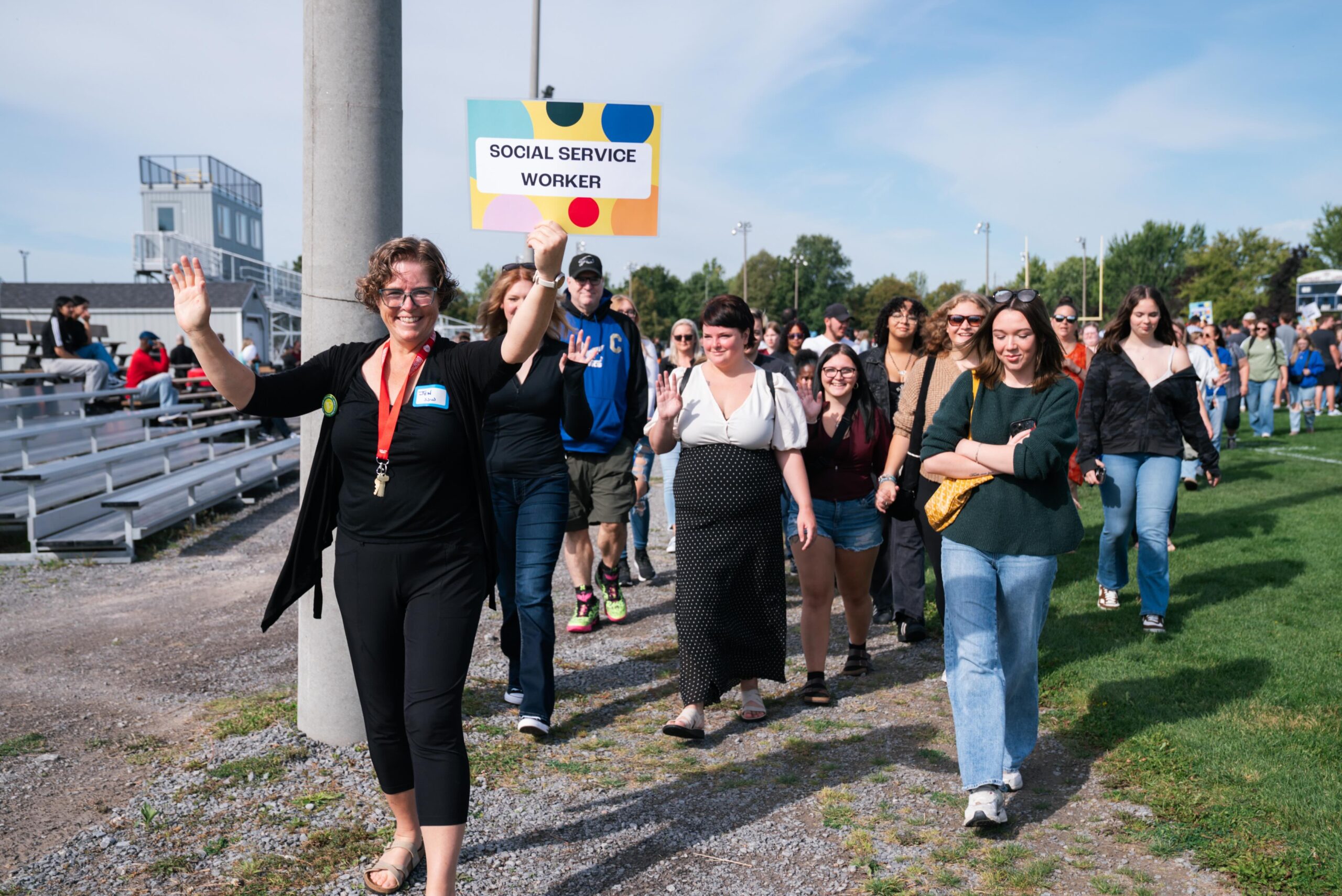 A group of Social Service Worker students walk together outdoors during an orientation event. A staff member at the front smiles and waves while holding a colourful sign that reads “Social Service Worker.” Students follow behind, also smiling and waving, with bleachers and a grassy field visible in the background.