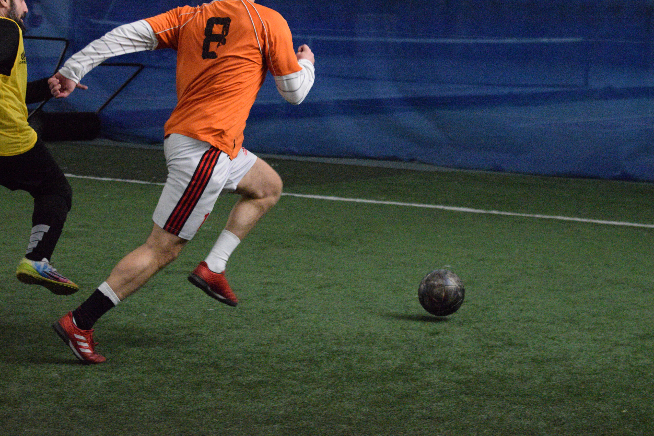 Two indoor soccer players compete for the ball on an artificial turf field, with one player in an orange jersey moving forward.
