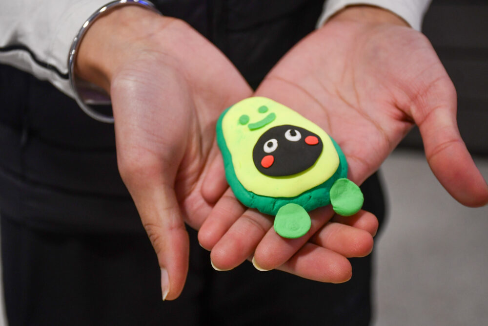 Two open hands holding a small handmade clay figure shaped like an avocado, with a smiling face and green details.