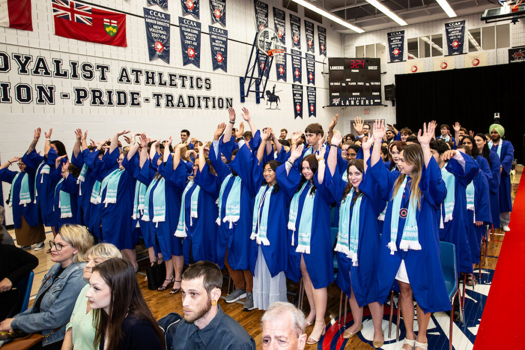 Loyalist College graduates raise their hands in celebration at an indoor convocation ceremony.