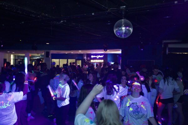 A crowded indoor dance party under blacklight, with a disco ball hanging from the ceiling and people wearing white T-shirts decorated with neon paint. Many are holding glowing light sticks and wearing glow necklaces, creating bright colours against the dark room as they dance and socialize.