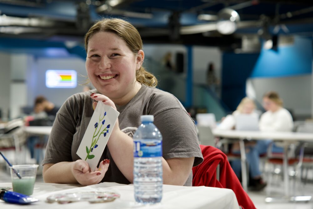 A smiling person sits at a table in a bright indoor space, holding up a small white card with a hand-painted design of blue flowers and green stems. A paintbrush and a cup of green paint sit on the table in front of them, along with a water bottle. Blurred people are seated at tables in the background.