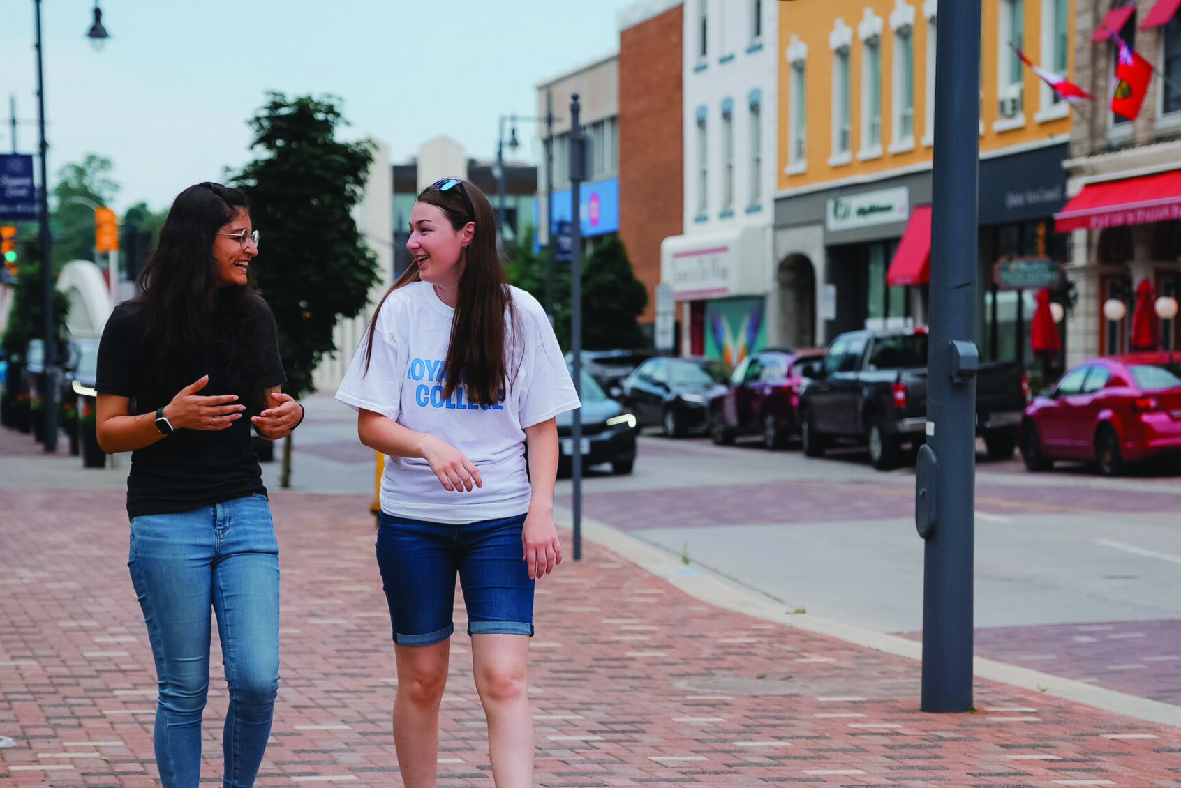 Two students walking and talking on a brick street in a downtown area, with one wearing a Loyalist College t-shirt and colourful storefronts in the background.