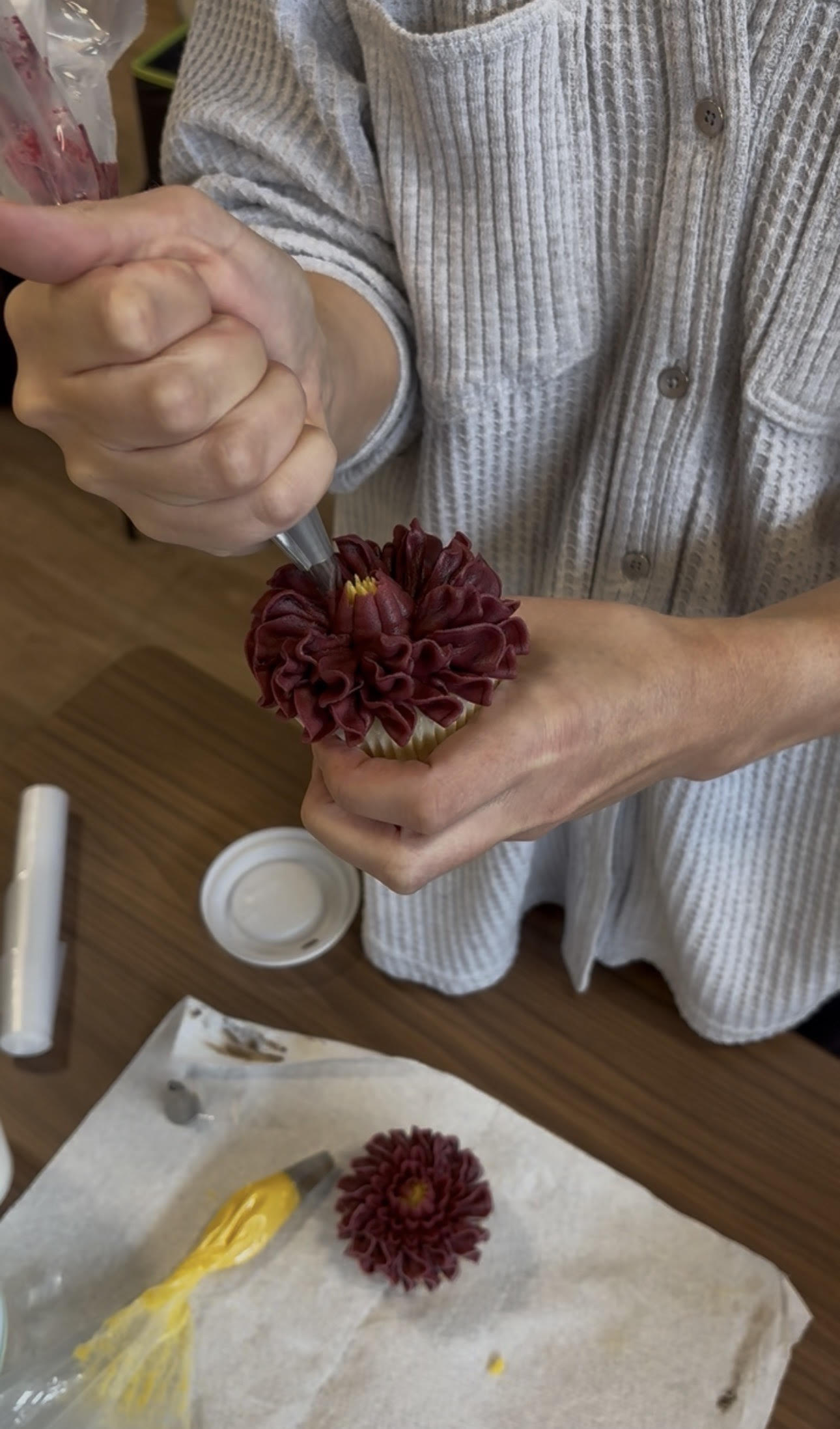 Close-up of a person wearing a light gray sweater piping dark red buttercream frosting onto a cupcake in layered, petal-like ruffles to form a flower with a yellow center. A second decorated cupcake, a yellow-filled piping bag, and baking tools rest on a wooden table below.