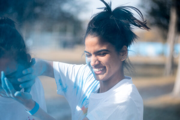 A woman outdoors smiles with her eyes closed as she playfully smears bright blue powder onto another person’s face. She is wearing a white T-shirt with blue powder on it and has her hair tied up. The background is blurred, suggesting an outdoor event or park setting.