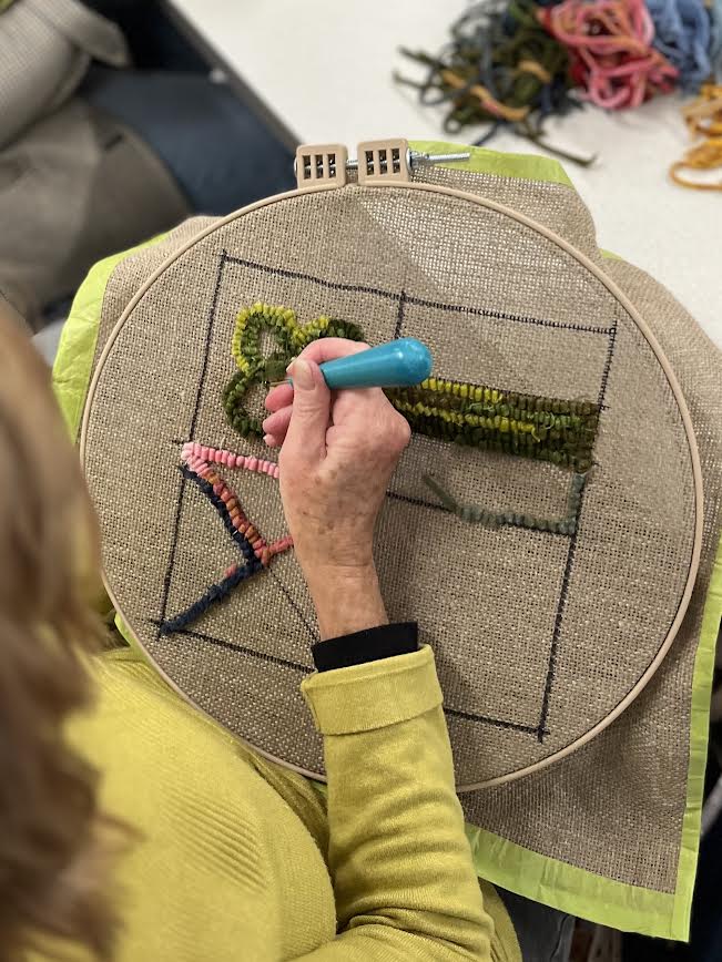 Overhead view of a person wearing a yellow top using a turquoise punch needle to stitch thick green yarn into fabric stretched in a round embroidery hoop, with a hand-drawn rectangular outline, colorful textured stitches, and loose yarn visible in the background.