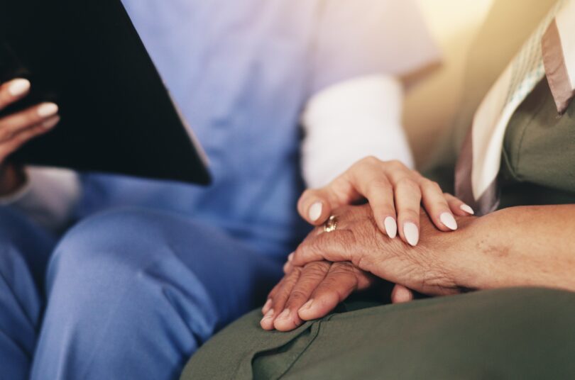 A healthcare professional in blue scrubs sits beside an older adult and gently holds their hands in a supportive gesture. The professional holds a tablet in one hand while the older adult rests their hands on their lap. The focus is on the hands, conveying comfort, care, and reassurance in a clinical or care setting.