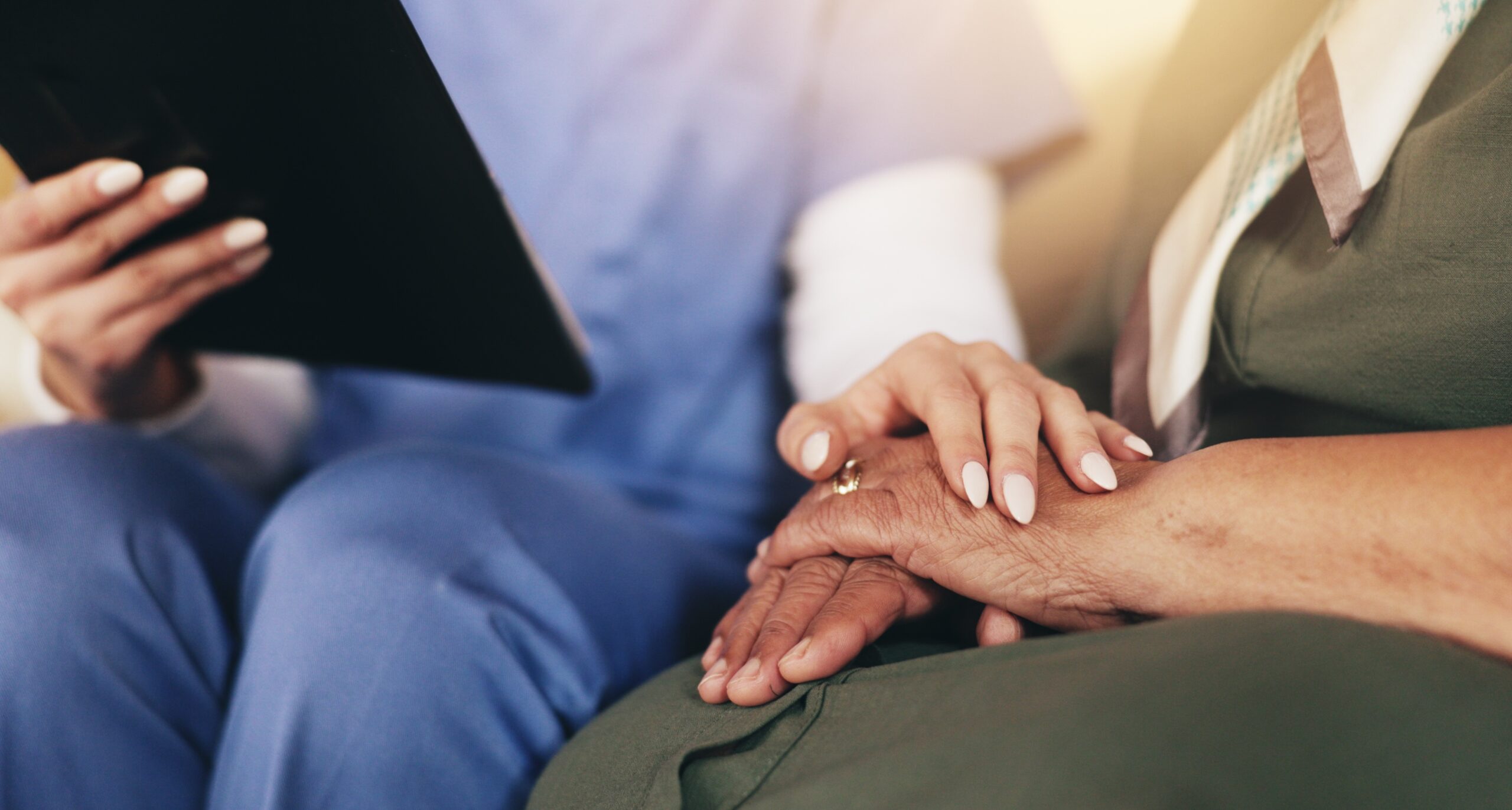 A healthcare professional in blue scrubs sits beside an older adult and gently holds their hands in a supportive gesture. The professional holds a tablet in one hand while the older adult rests their hands on their lap. The focus is on the hands, conveying comfort, care, and reassurance in a clinical or care setting.