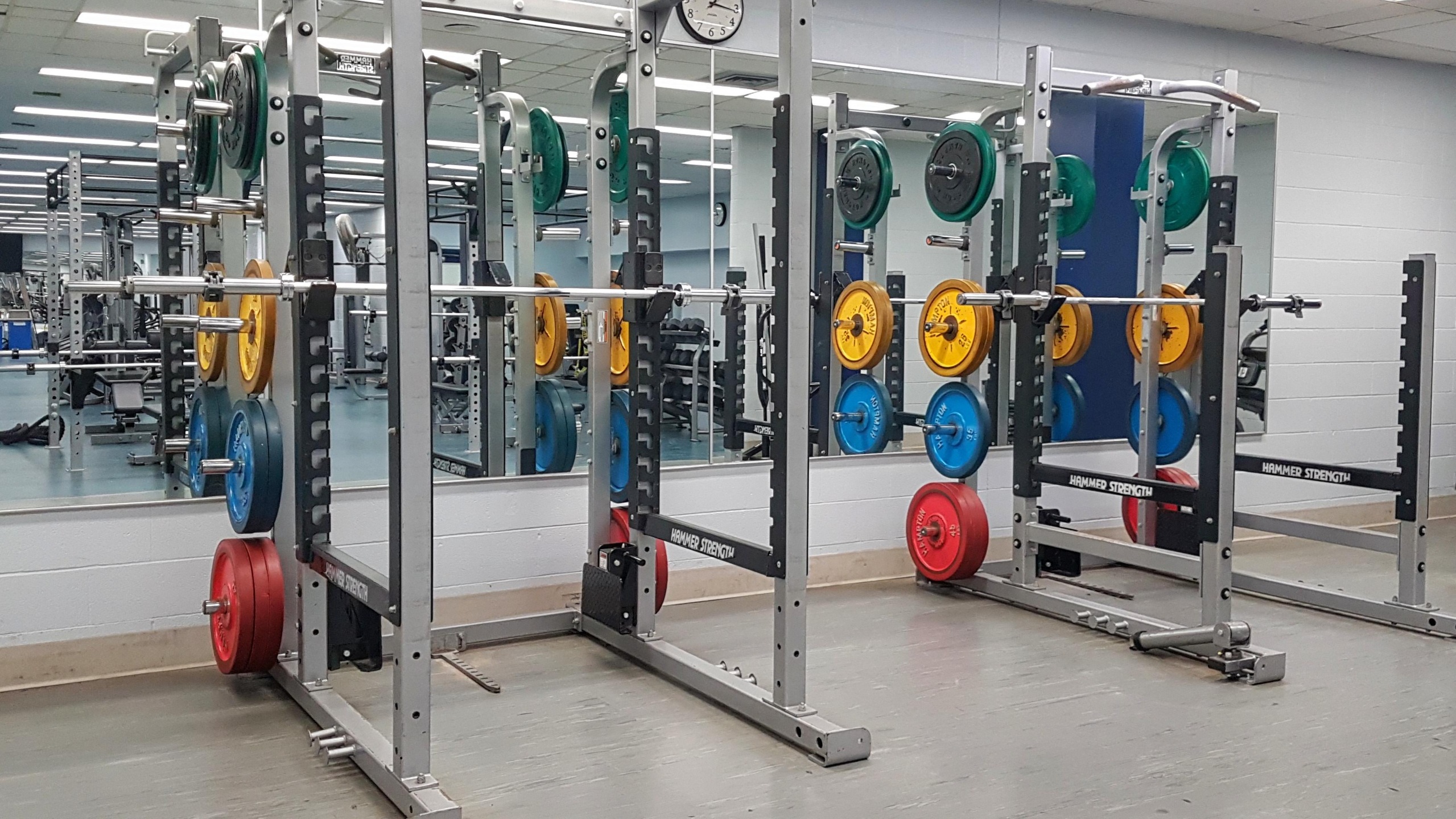Two Hammer Strength power racks loaded with coloured bumper plates in red, blue, yellow, and green, in a fitness centre with mirrored walls and additional weight equipment visible in the background.