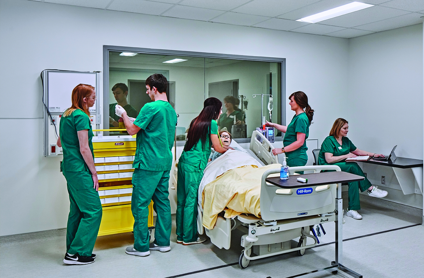 Students in scrubs practicing patient care in a simulated hospital room, working together around a patient bed with medical equipment.