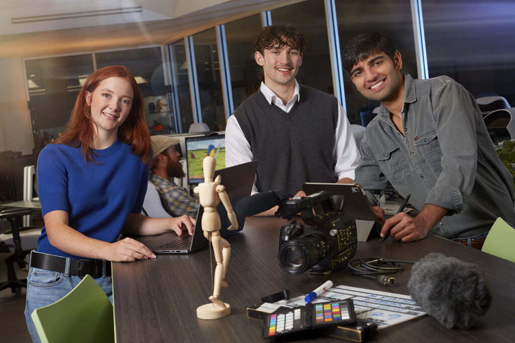 Three students collaborating on a media project with cameras, laptops and audio equipment in a creative studio setting.