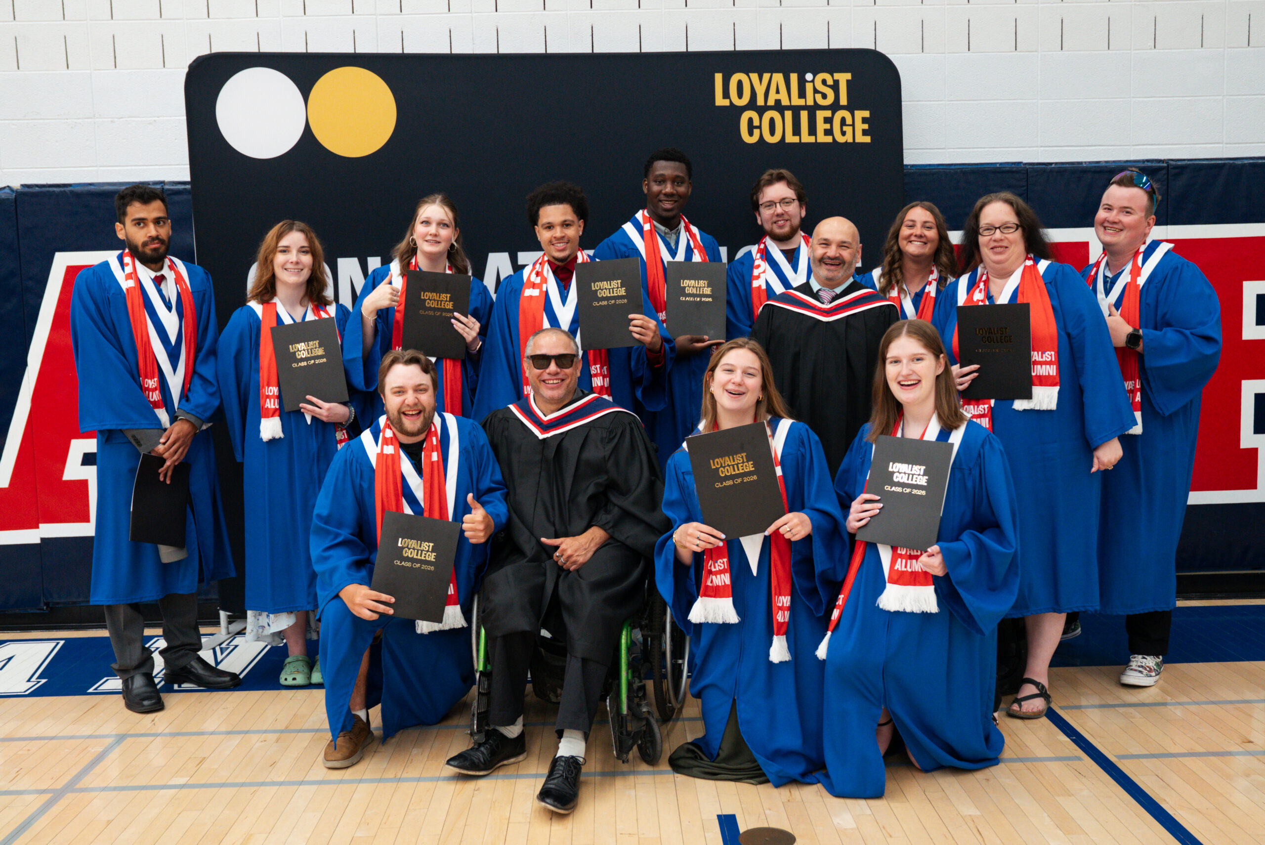 A large group of Loyalist College graduates and a faculty member in academic regalia pose together in front of a Loyalist College backdrop, holding up their diplomas.