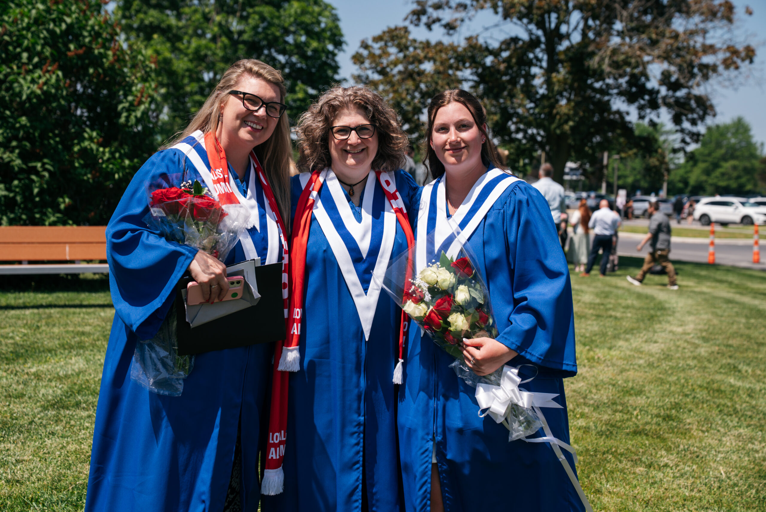 Three graduates in blue convocation gowns and white stoles smile together outdoors, two holding bouquets of flowers, at a graduation ceremony.