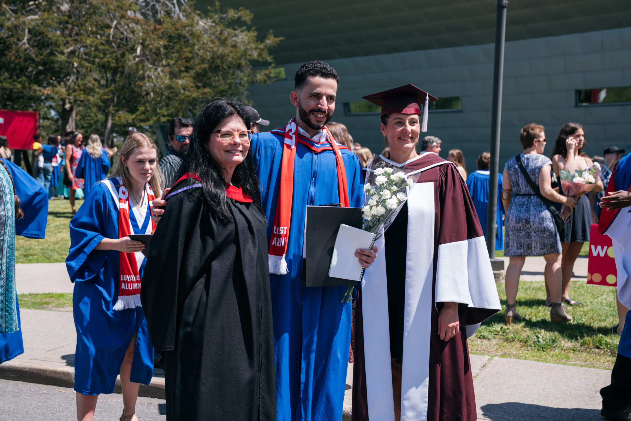 Three people in academic regalia smile together outdoors at a convocation ceremony, with a crowd of graduates and guests visible in the background