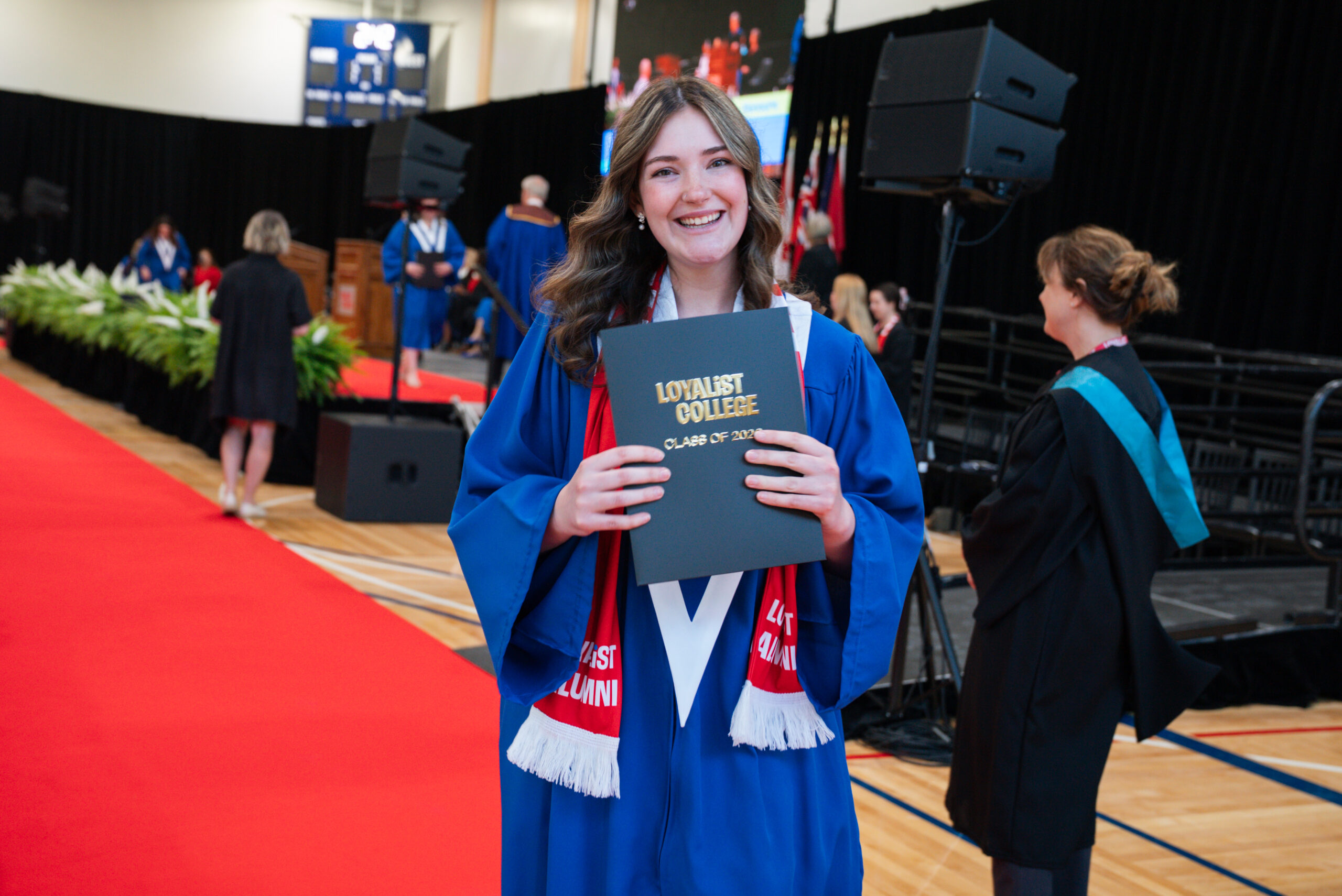 A smiling graduate in a blue convocation gown holds up a Loyalist College Class of 2026 diploma folder inside the convocation ceremony venue.