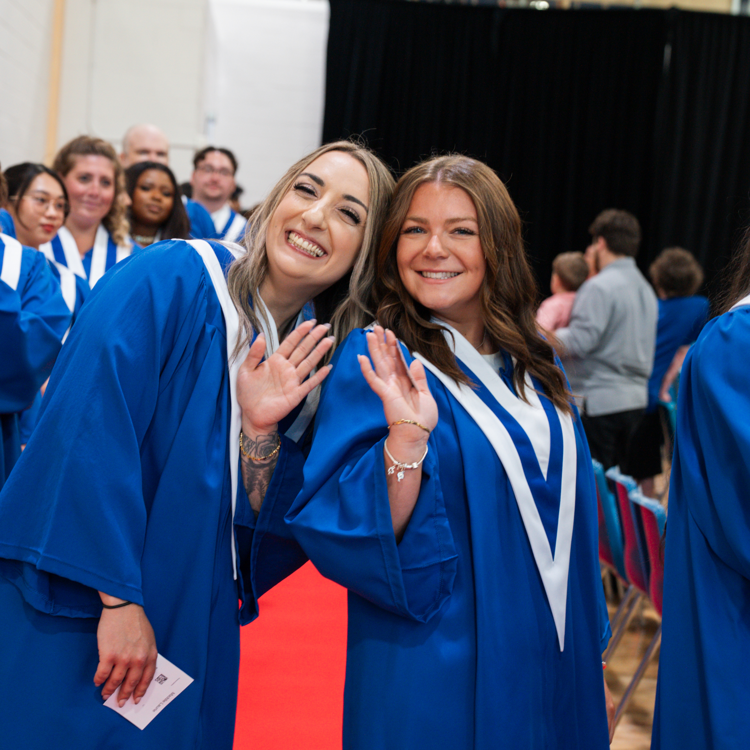 Two graduates in blue convocation gowns wave and smile at the camera inside the convocation venue, with fellow graduates visible in the background.