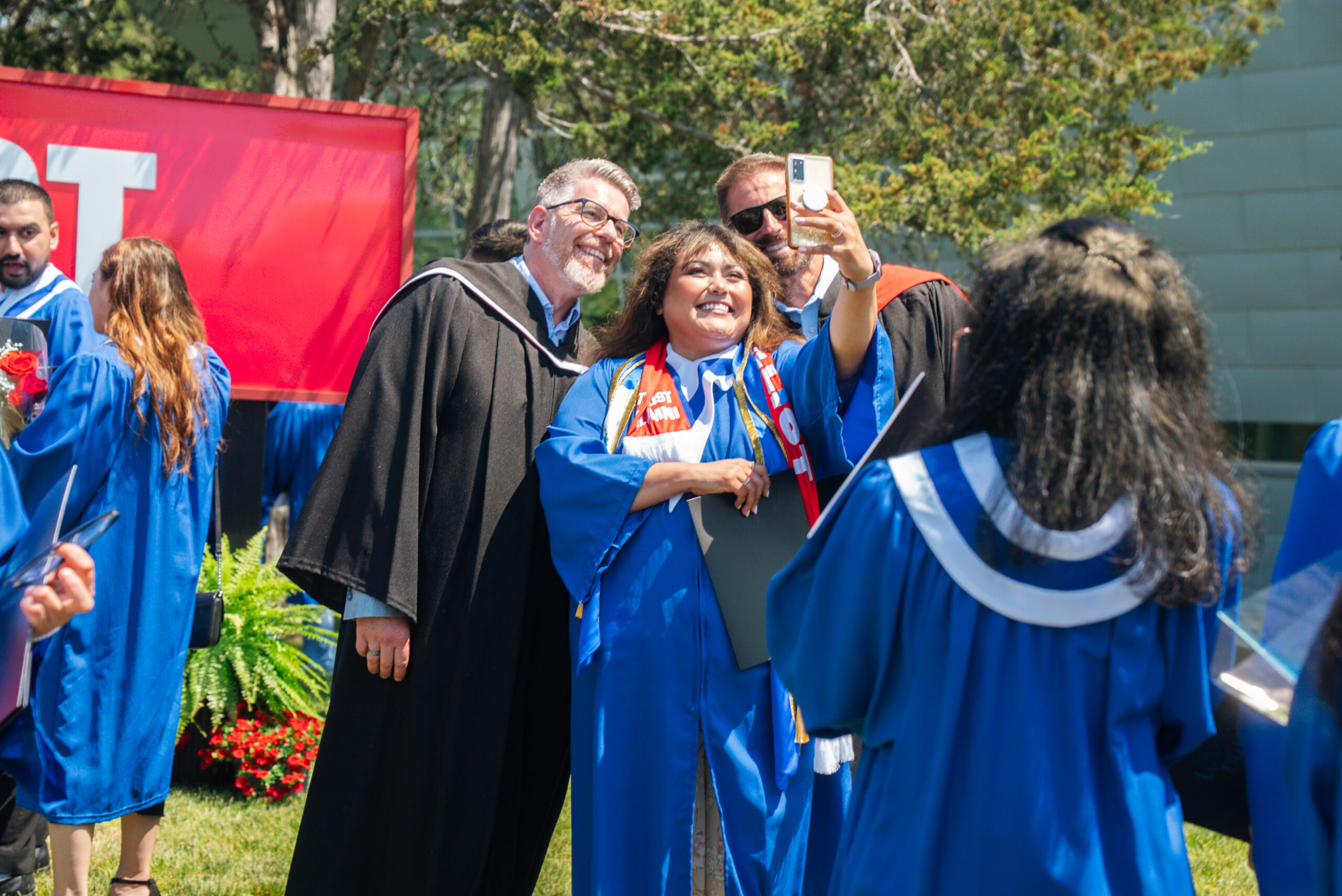 A graduate in a blue convocation gown takes a selfie with two people in academic regalia outdoors on a sunny convocation day.