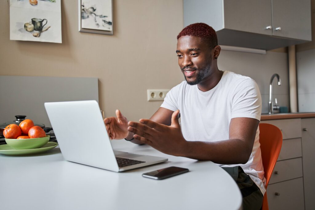 A man sitting at a kitchen table participating in a video call on a laptop, gesturing while speaking.