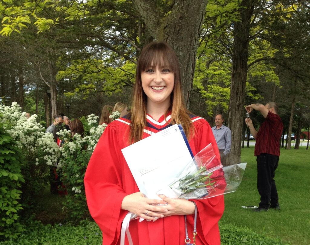 A female graduate in a red gown holds a diploma and a bouquet outdoors.
